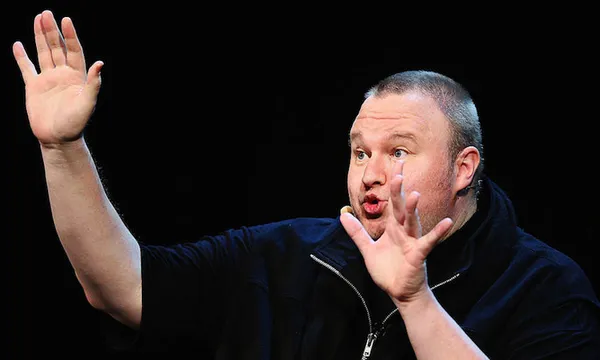 a tall whit man with his lips pursed and ams out looking dramatic, wearing a zip up black polo shirt on a black background