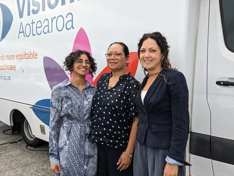 three women wearing blue tones outside a coloursful bus. Sachi, on the levt is short, with light brown skin, tightly curling hair and a big smile; Sila, in the middle is a pacific woman with straight hair tied back and rectangular glasses. Germaine, on the right, is a lebanese woman with no glasses and a friendly smile