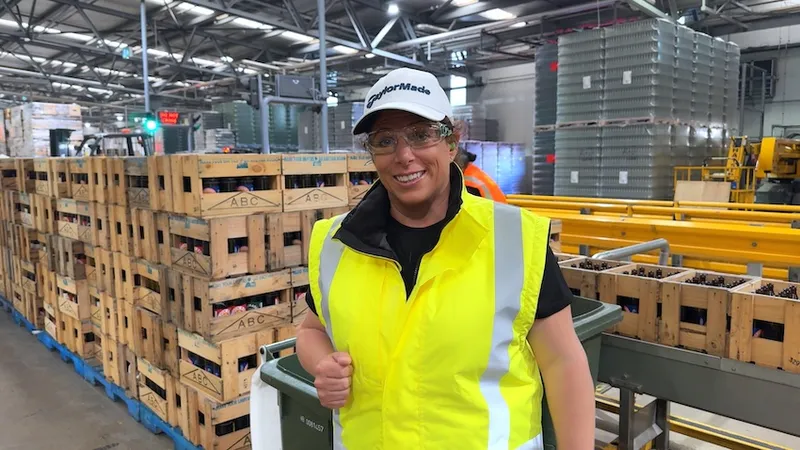 a fun and down to earth looking woman with short hair and a big smile and hi vis fest wears a cap inside a waste processing facility of some kind, with lots of cartons behind her