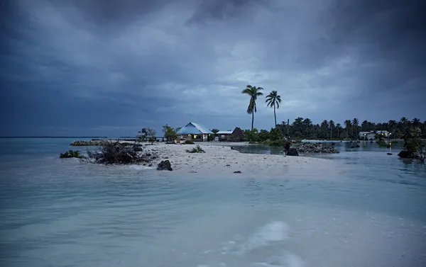 a stormy purple sky, with some houses on a pale islane and a pale turquoise swish of water in the foreground