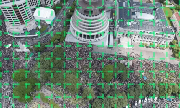 A large crowd gathers outside a circular government building, surrounded by trees and other structures, seen from above. Green grid overlays the image.