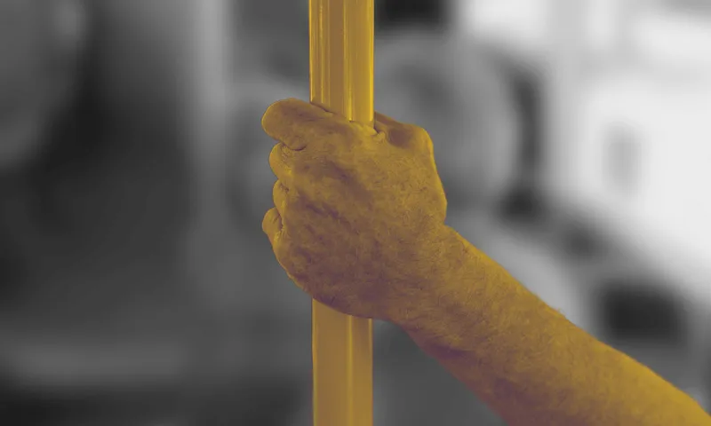 A close-up of a person's hand holding onto a vertical pole inside a public transportation vehicle. The image is rendered in a yellow monochrome filter, with the background slightly blurred.