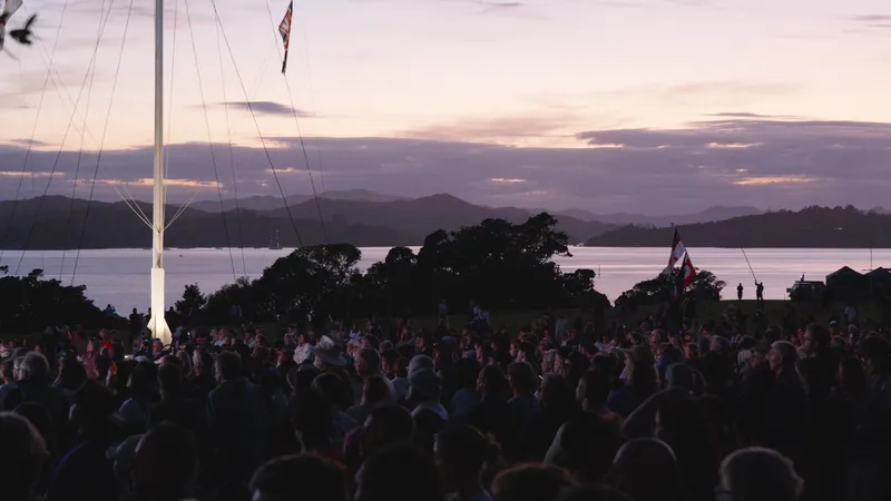 A darkened shot at sunrise, with a crowd of people gathered on the upper Treaty Grounds at Waitangi, watching a big screen, not pictured