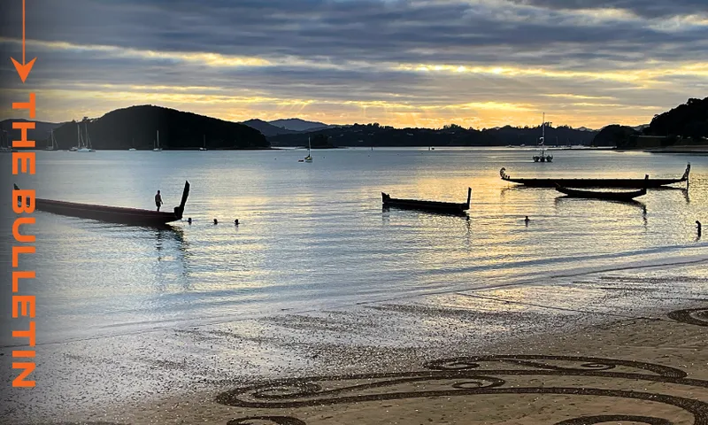 Waka at Waitangi, February 6, 2025 (Photo: Anna Rawhiti-Connell) Serene beach scene at dawn at Waitangi with several waka silhouetted on calm water. Gentle waves lap the shore, which features intricate sand art in the foreground. Text on the left reads "The Bulletin".