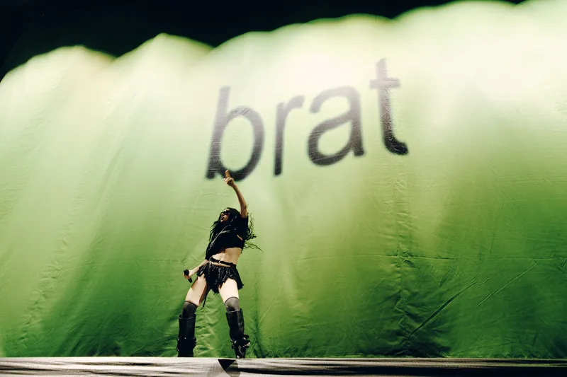 Charli XCX in front of a bright greet banner with "brat"
