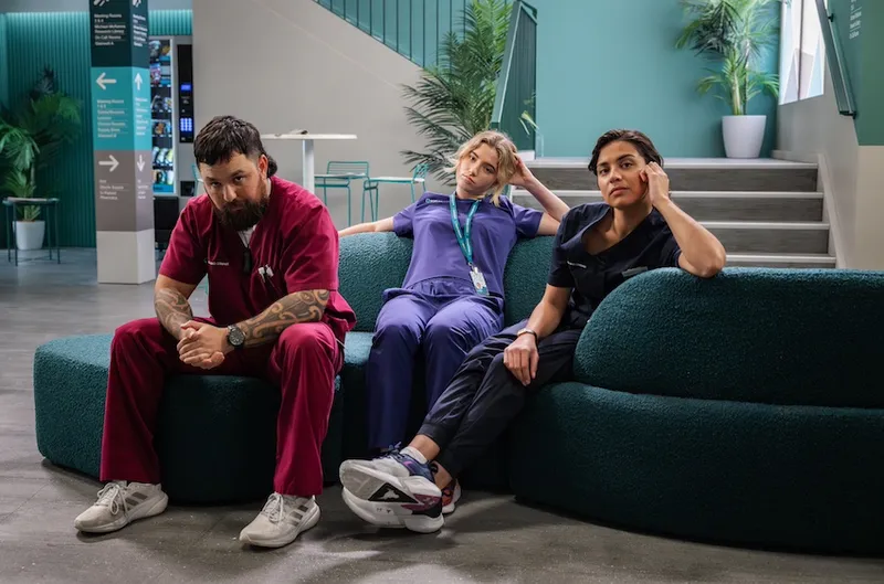 Three people wearing medical scrubs sit on a dark green couch and stare at the camera