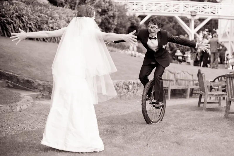 Ken unicycles towards Anna at their wedding. He is wearing a black tuxedo, she is wearing a white dress. They both have their arms outstretched.