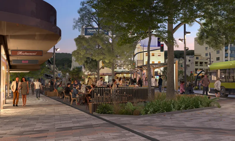 A vibrant urban street scene at dusk with people dining at outdoor cafés, surrounded by trees and modern shops. A green bus is visible to the right. Warm lights illuminate the area, creating a welcoming atmosphere.