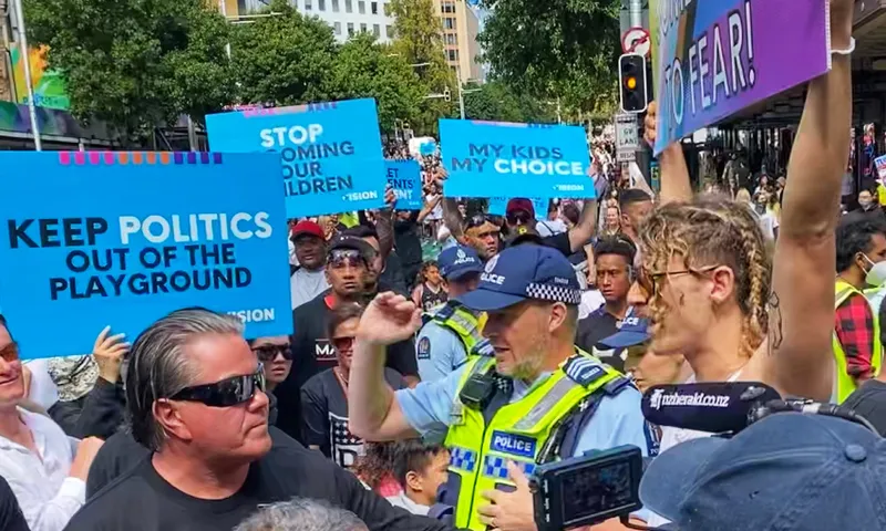 A protest with people holding signs such as "Keep politics out of the playground" and "My kids, my choice." A police officer talks to Brian Tamaki. The crowd is diverse, with trees and buildings in the background.