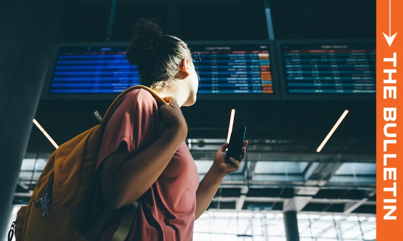 A person with a backpack looks up at flight information screens in an airport terminal. They are holding a phone. Sunlight streams through the large windows. An orange vertical banner on the right reads "The Bulletin.