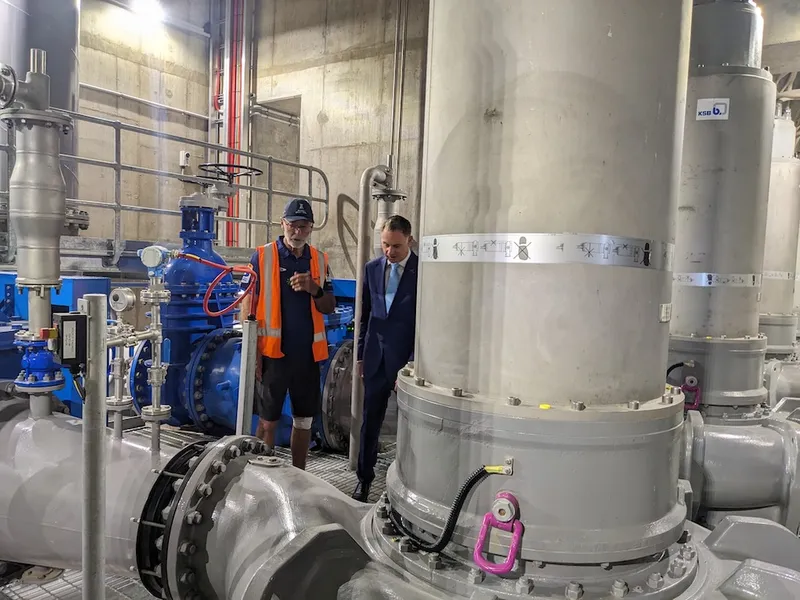 two white men, wane brown in a tshirt and hi vis and simon wats in a suit, walk among huge pipes