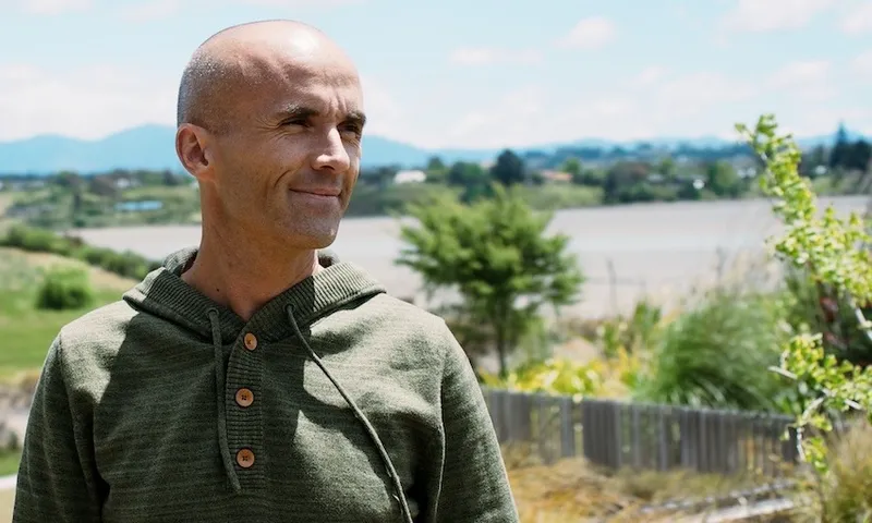 a bald man with plants in the backgroun and a buttoned green hoodie