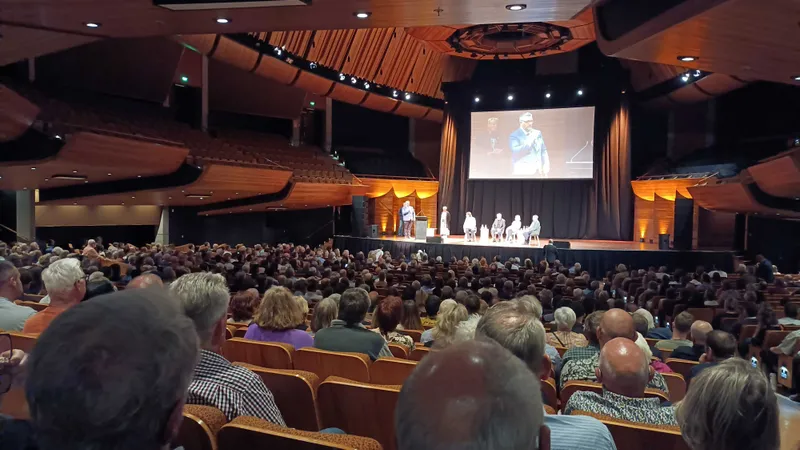 A photo taken at the back of a crowd of people, many with grey hair, watching four panellists on stage