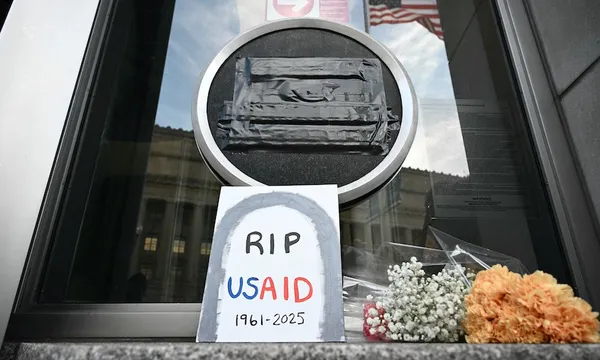 A window with a taped circular emblem displays a sign resembling a gravestone that reads, "RIP USAID 1961-2025." Below it, there are a bouquet of white flowers and yellow flowers. The American flag is reflected faintly in the window.