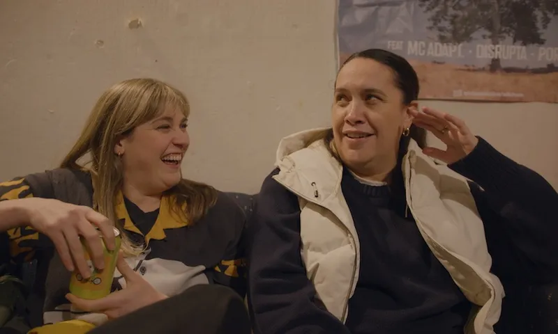 brynley and kura laughing on a couch in a sparsely decorated student flat. behind them is a poster for a drum and bass gig