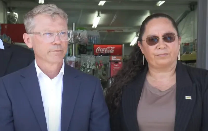 Two individuals stand in front of a store entrance. The person on the left, justice minister Paul Goldsmith, is wearing a dark suit and glasses, and the person on the right, associate justice minister Nicole McKee, is wearing a striped blazer with a pin. Both appear serious, with a Coca-Cola sign visible in the background.