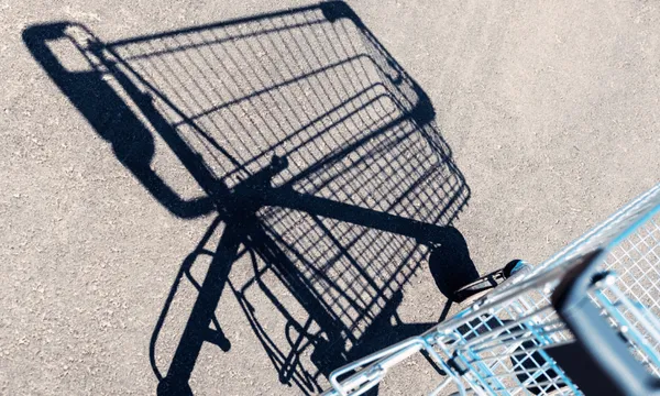Shadow of an empty shopping cart cast on a sunlit concrete surface, with the cart's metallic structure partially visible in the bottom right corner.