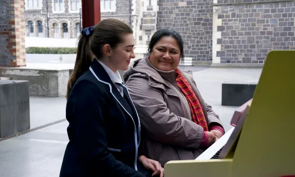 Sophie, a young white woman in a school uniform sits at a piano next to Mareta, an older Samoan woman wearing a coat and scarf