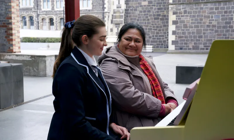 Sophie, a young white woman in a school uniform sits at a piano next to Mareta, an older Samoan woman wearing a coat and scarf