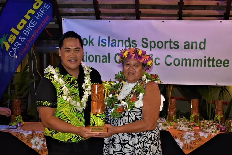 A man and a woman, both adorned with floral garlands, stand together smiling. The man holds a trophy. A banner behind them reads "Cook Islands Sports and National Olympic Committee." Tables with trophies and floral decorations are in the background.
