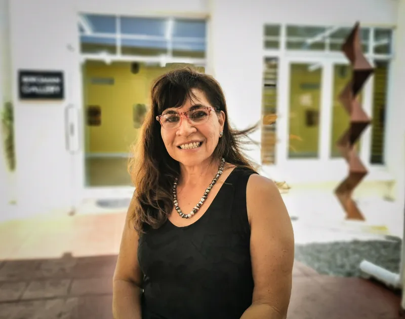 A smiling woman with long, dark hair and glasses stands outside a building with large windows. She is wearing a black sleeveless top and a beaded necklace. A tall, abstract sculpture is visible in the background.