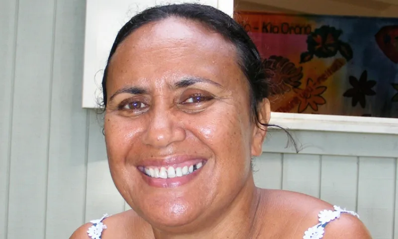 A person smiling while sitting in front of a wall with a colourful floral painting. The lighting is natural, and the person is wearing a white top.