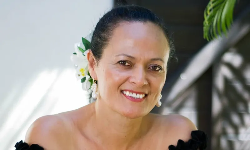 A person with a flower in their hair smiles warmly, wearing off-shoulder black attire and pearl earrings. They're standing outdoors, with greenery and natural light in the background.