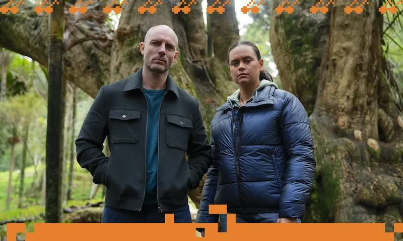 A man wearing a black jacket and a woman wearing a blue down jacket stand closely to each other in a forest and look directly at the camera
