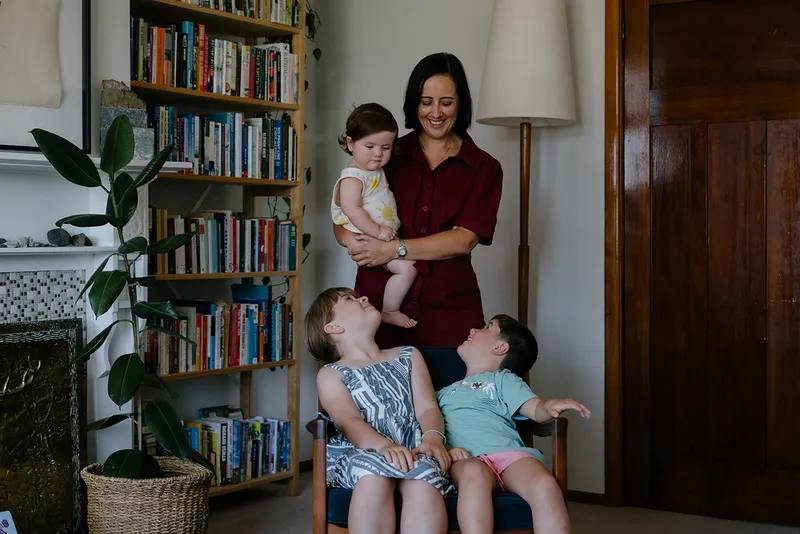 photo of woman in a red dress holding a baby with two other small children. bookshelf behind them