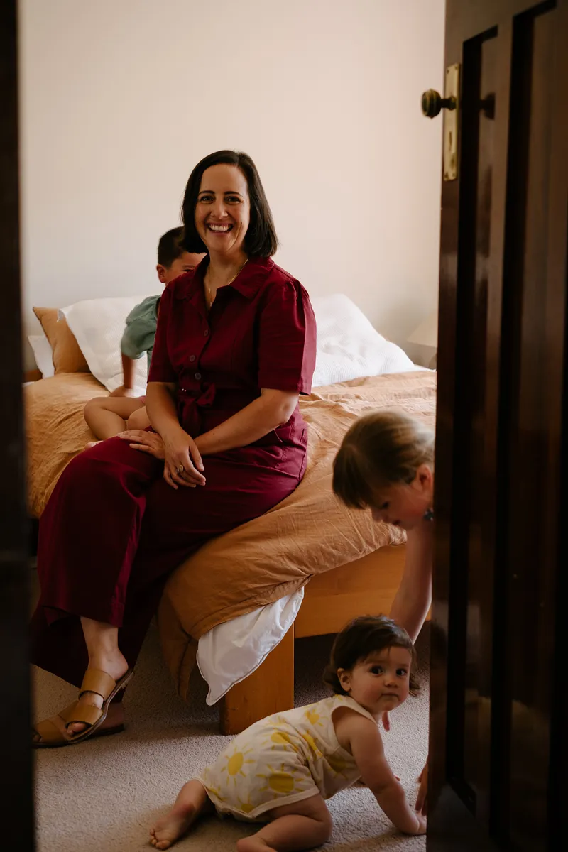 woman in red jumpsuit on a bed with three children playing around her