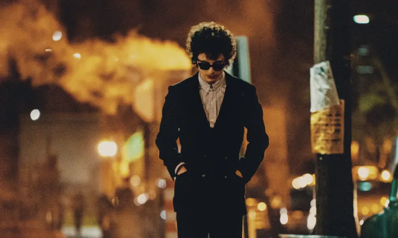 A person (Timothée Chalamet as Bob Dylan) with curly hair and wearing sunglasses walks down a dimly lit street at night. They are dressed in a dark suit with hands in pockets. Bright lights and smoky air create a moody atmosphere in the background.