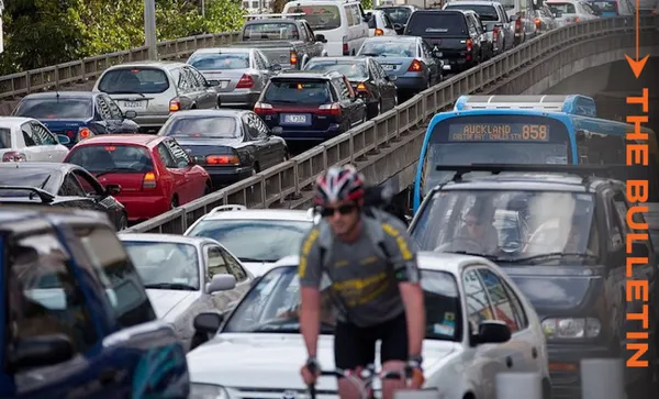 Heavy traffic on a multi-lane road with numerous cars and a blue bus. In the foreground, a cyclist in a helmet and sunglasses rides along the congested road. The words "The Bulletin" are on the right side in orange text.