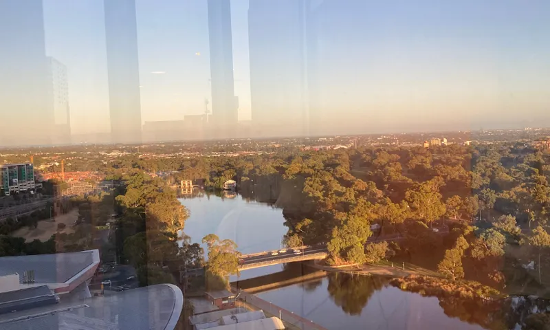 Aerial view of a cityscape at sunset. A river runs through a park with dense trees, crossed by a bridge. Buildings and urban infrastructure are visible in the background, reflecting warm sunlight and soft, clear skies above.