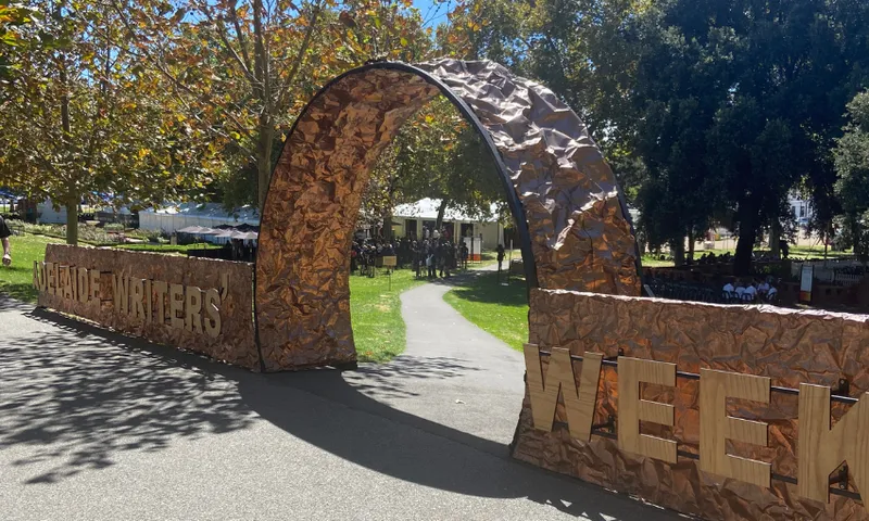 The image shows an outdoor entrance archway with crumpled, metallic-looking decorations and large letters spelling "ADELAIDE WRITERS WEEK." There are trees and a grassy area with tents in the background.