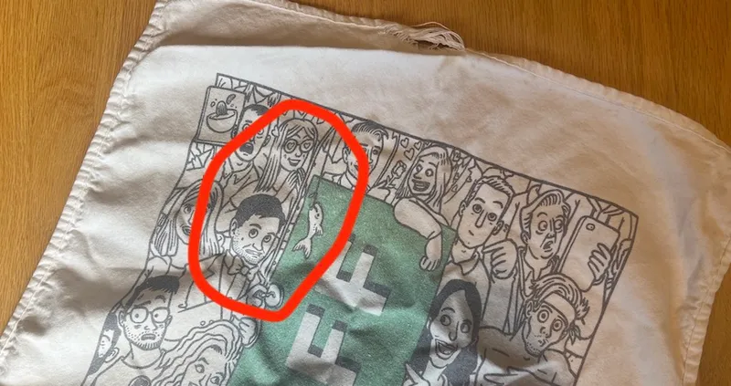 Illustration on fabric showing a crowd of cartoon characters, including one circled in red. The characters have varied expressions and are holding smartphones. The fabric appears worn at the top edge.