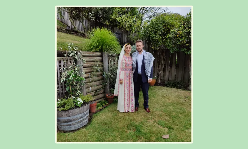 a couple in the back garden with green grass. Noor, in a white embroidered dress, stands beside matt, in a suit with a keffiyeh around his shoulders