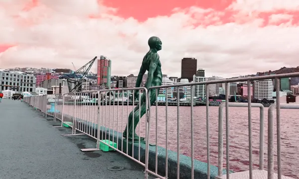 A bronze sculpture of a man appears to be diving into the water, positioned on a waterfront with railings. Urban buildings and construction cranes are visible across the water under a cloud-filled sky with a pinkish hue.