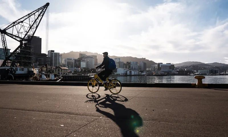 Person riding a yellow bike on a waterfront promenade with city buildings and hills in the background. The sun is shining brightly, casting a long shadow. An industrial crane stands to the left. The scene is set against a partly cloudy sky.
