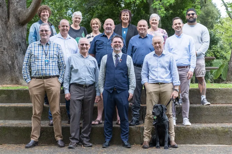 A group of 14 people pose on outdoor steps, smiling at the camera. One person has a black dog. They are in a park with trees in the background. The group includes a mix of men and women in casual and business casual clothing.