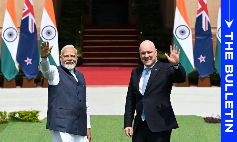 Two men in formal attire wave while standing on a lawn. Behind them are Indian and New Zealand flags. The setting is outdoors with a red carpet leading to a building.