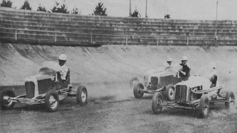 Three vintage race cars, each with a driver wearing a helmet, speed around a dirt track on a banked curve. The grandstand in the background is empty, and trees are visible behind the seating area. Dust trails behind the cars, adding a dynamic feel.
