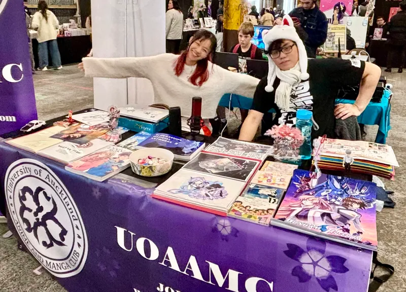 two anime fans sit behind a stall at a convention with anime comics in front of them