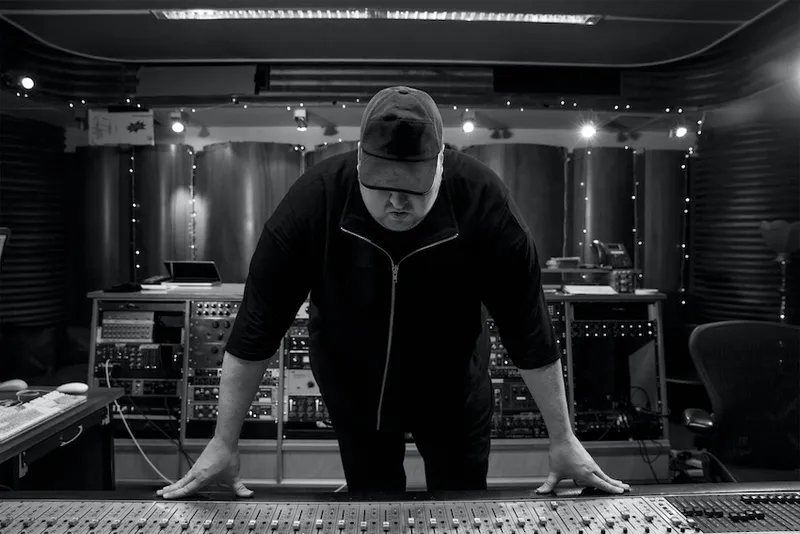 A person in a cap leans over a mixing console in a dimly lit recording studio. The space is filled with audio equipment and illuminated by soft lights. The person is focused on the controls and wears a dark jacket.