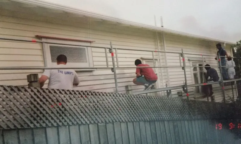 an old film photo of five adults painting the side of a house while on a large scaffolding