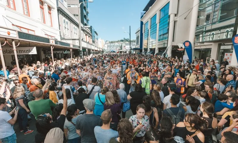 A large crowd gathers on a city street, celebrating with music and dancing. Bright buildings line the street, and the sky is clear. People are dressed casually, some holding cameras and phones, capturing the lively atmosphere.