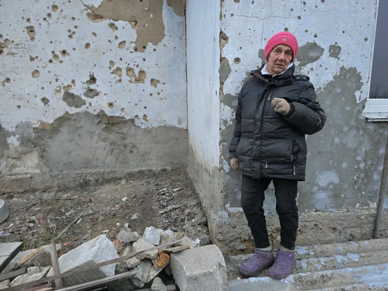 A woman in a pink beanie and puffer jacket stands in front of a home riddled with bullet holes