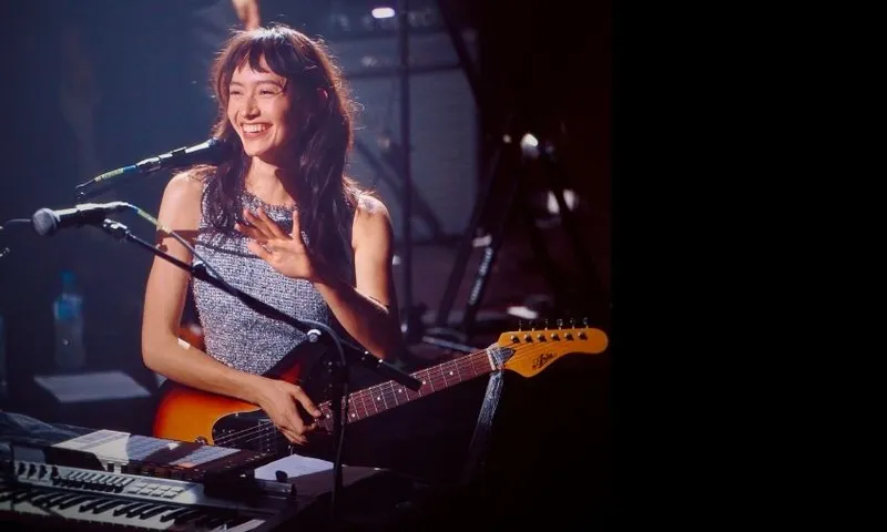 Amelia Murray Fazerdaze smiling on stage with an electric guitar and microphone, wearing a sleeveless top. She stands near a keyboard, with stage lights and musical equipment visible in the background.