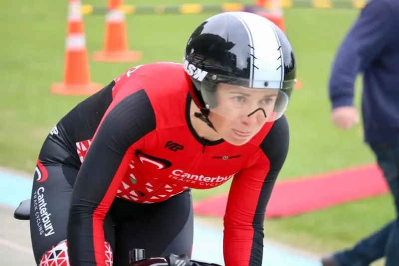 Fox, a person with pale skin and a focused expression wears red and black lycra and a black helmet, loking fast