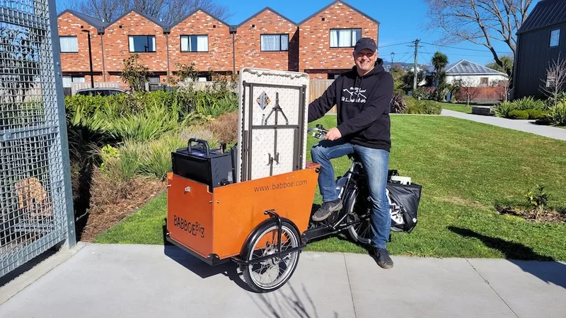 a white man with jeans smiling on a cargo bike with lots of stuff in the cart