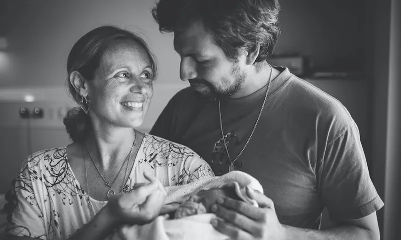 A couple lovingly looks at each other while holding a newborn wrapped in a blanket. Both are smiling, standing closely, with soft lighting creating an intimate atmosphere. The image is in black and white.